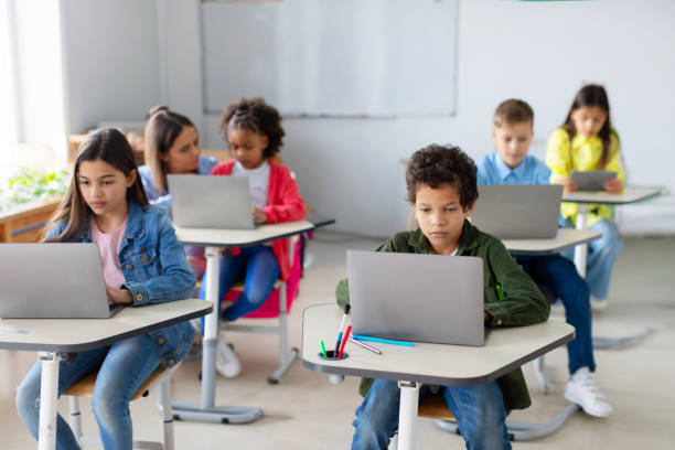 Diverse school children sitting at desks with laptops, studying using digital gadgets. Modern elementary education concept